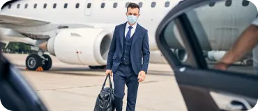 A man in a blue suit and face mask carrying a leather bag walks from a private jet toward a waiting luxury car.
