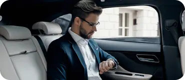 A businessman in a navy suit sits in the back seat of a white-leather luxury car interior, looking at his wristwatch